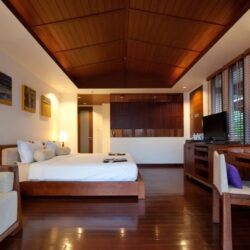 Interior view of The Sarann Koh Samui’s spacious deluxe room with a king-size wooden bed, side lamps, seating area and large windows with wooden blinds.