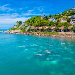 The turquoise ocean leads up to the Sarann Koh Samui beach resort, nestled amongst a rocky coast backed by tropical trees and hillside accommodation under a bright blue Thailand sky.