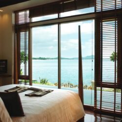 The Sarann Koh Samui's Sea Front Pool Villa Seaview features a white-lined bed, floor-to-ceiling windows with wooden blinds, and a view of a private pool and the turquoise sea horizon.