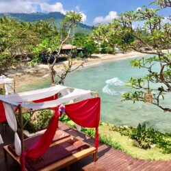 Elevated sea view from The Sarann Koh Samui with draped sunbeds on a wooden platform, framed by flowering trees and lush tropical greenery.